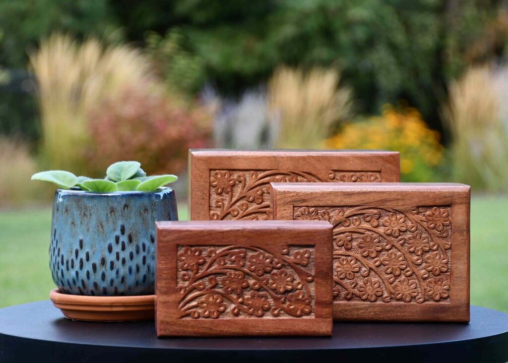 Set of three acacia wood urns with intricate floral carvings, an elegant upgrade option from Furevergreen Aquamation, displayed beside a blue ceramic planter with green leaves.